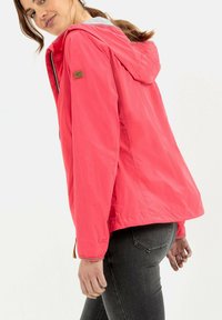 Veste à capuche rose vif en matériau léger, avec fermeture éclair, poignets élastiques et un écusson logo sur la manche.