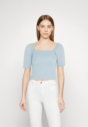 Svetle modrý crop top se čtvercovým výstřihem, krátkými nafouknutými rukávy s květinovou výšivkou, spojený s bílými džíny s roztrhnutím.