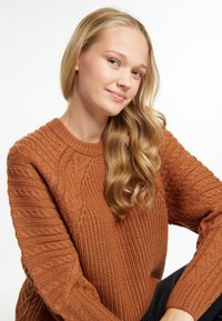 Gruby pomarańczowy sweter z dzianiny o fakturowych wzorach, w tym splotach kablowych i ściągaczach, okrągły dekolt i długie rękawy. Gładkie tło.