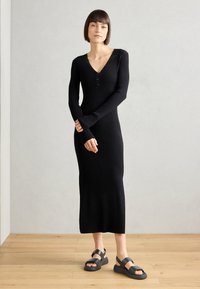 Black ribbed knit dress with a V-neckline and button detail, long sleeves extending past wrists, worn with black sturdy sandals.
