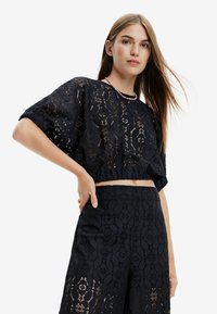 Sort cropped top med blonder og blomsterdesign, rund halsudskæring og korte pufærmer, parret med matchende blondebukser.