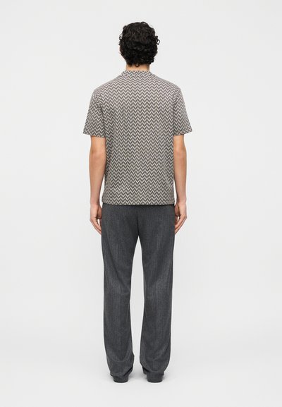 Άνδρας με σγουρά μαλλιά στέκεται με την πλάτη γυρισμένη, φορώντας κοντομάνικο πουκάμισο με ζιγκ-ζαγκ σχέδιο και φαρδιά σκούρα γκρι παντελόνια σε ένα απλό φόντο.