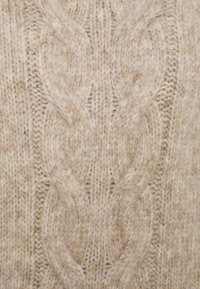 Beiger gestrickter Pulloverstoff mit einem strukturierten Kabelmuster aus weichem, flauschigem Garn. Sichtbare Nähte und unterschiedlich dickes Garn.