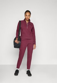 Costum sport burgundiu compus dintr-un hanorac cu fermoar și pantaloni cargo. Material texturat cu buzunare; asortat cu adidași negri și o geantă.