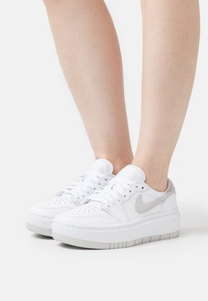 Zapatillas de cuero blanco con punta perforada, acentos en gris y una suela blanca gruesa. Cuerdas detalladas y logo distintivo en el lateral.
