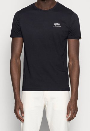 Czarny krótki rękaw T-shirt Alpha Industries z małym logo na lewej piersi, noszony przez osobę o ciemnej karnacji, ubrana w białe spodnie.