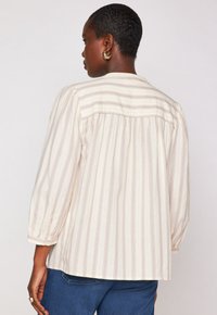 Persona di colore scuro con capelli corti, che indossa orecchini a cerchio dorati, una blusa a righe crema e beige con maniche a 3/4 e jeans blu, di spalle.