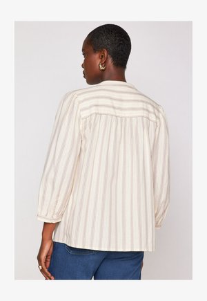 Persona di colore scuro con capelli corti, che indossa orecchini a cerchio dorati, una blusa a righe crema e beige con maniche a 3/4 e jeans blu, di spalle.