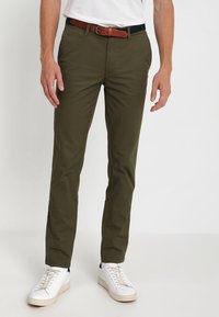 Olivgröna chinos i bomull med en slät struktur, utrustade med ett brunt läderbälte och vita sneakers med krämfärgade sulor.