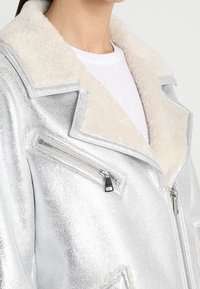 Giacca metallizzata argento con colletto in peluche di colore crema. Presenta una tasca sul petto con zip e un materiale testurizzato con un design elegante.