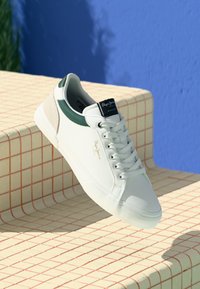 Zapatilla blanca con detalles en verde y beige, parte superior de cuero suave, puntera redonda y suela plana de goma. Presenta un acabado texturizado y el logotipo de la marca.