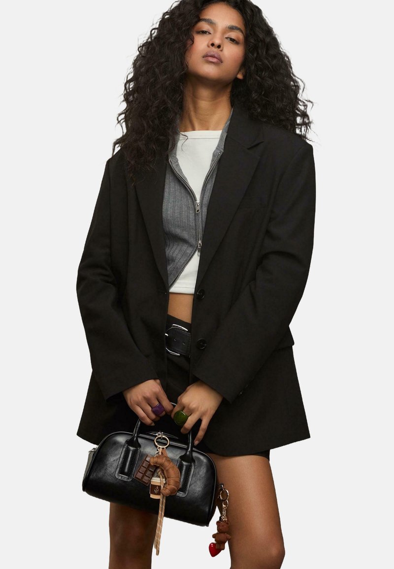 Ung kvinde med krøllet hår iført en lagdelt sort blazer og en cropped top, som holder en sort håndtaske prydet med nøgleringscharms.