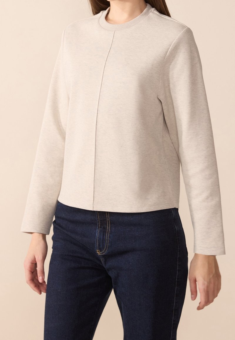 Sweat-shirt gris clair avec col rond, manches longues et coupe droite. Présente une couture centrale comme détail de design. Porté avec un jean foncé.