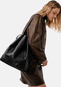 Donna con giacca di pelle marrone e gonna, che porta una grande borsa a tracolla in pelle nera con texture, guarda verso il basso.