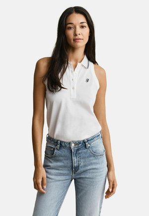 Femme aux longs cheveux noirs portant un polo blanc sans manches avec un petit logo et un jean taille haute bleu clair sur un fond blanc.
