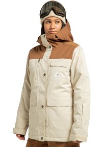 Beżowa wodoodporna kurtka z brązowymi ramionami, zamkiem błyskawicznym i patkami na kieszeniach, kapturem oraz naszywką z logo na piersi. Miękka w dotyku i ocieplana.