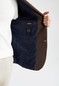 Mano che tiene aperto un blazer marrone con fodera interna blu scuro e un'etichetta che legge "STUDIO DeFacto SLIM FIT."