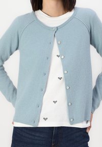 Himmelblaues Cardigan aus weichem Material, mit Rundhalsausschnitt, durchgehendem Knopfverschluss und gerippten Bündchen. Über einem weißen Hemd mit schwarzen Mustern getragen.