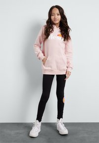 Sweat à capuche rose avec logo blanc, poche kangourou, porté sur des leggings noirs et des baskets blanches. Tissu lisse, coupe décontractée, style casual.