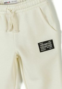 Pantalon pour enfants couleur crème avec taille élastique, cordon de serrage, poches latérales et un patch noir portant l'inscription "Unlimited Collective" sur la jambe droite.