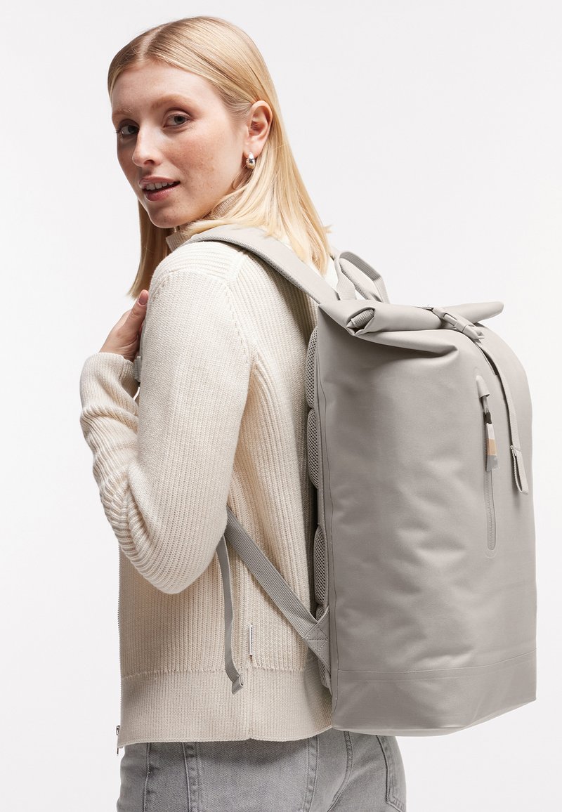 Jeune femme portant un pull léger et un jean, porte un grand sac à dos gris à fermeture roulante sur l'épaule sur fond blanc.