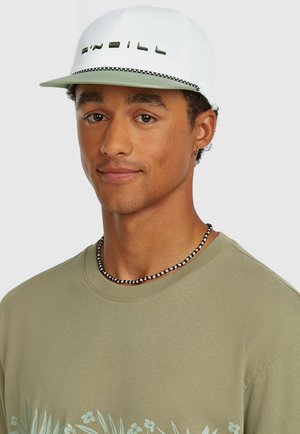 Valkoinen lippalakki, jossa vihreät korostukset ja etuosassa kirjailtu teksti sekä kontrastivärinen lippa. Malli käyttää vaaleanvihreää kukkakuviollista paitaa.