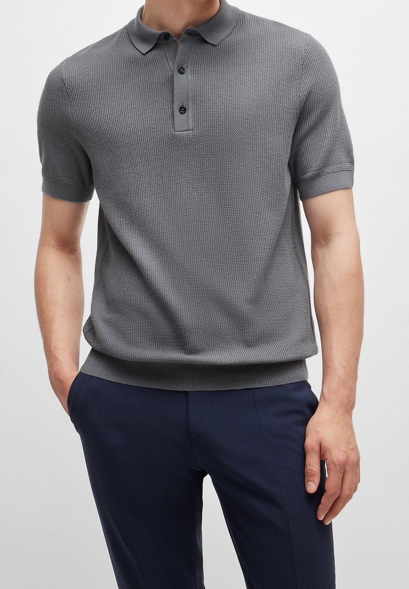 Homme portant un polo gris texturé à manches courtes et un pantalon bleu foncé, debout les mains dans les poches devant un fond uni.