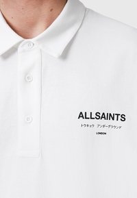 Hvit polo skjorte laget av bomull, med klassisk krage, tre knapper og svart grafisk tekst "ALLSAINTS" med japanske tegn og "LONDON."