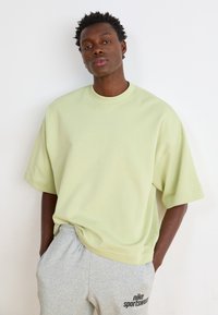 Világos zöld, oversized póló rövid ujjakkal, szürke melegítőnadrággal párosítva, amelyen fekete "nike sportswear" felirat található. Simább anyagtextúra.