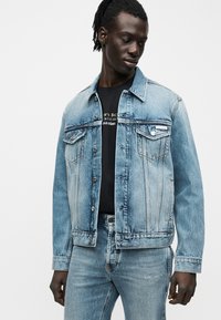 Άνδρας που φορά ανοιχτό μπλε τζιν μπουφάν και τζιν παντελόνι με μαύρο πουκάμισο Calvin Klein, κοιτάζοντας προς τα κάτω αριστερά σε ένα απλό φόντο.