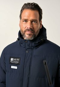 Marineblaues Steppjackett mit hohem Kragen, Frontreißverschluss und zwei Reißverschlusstaschen. Mit schwarzen Patches, die das Branding und besondere Texturdetails zeigen.
