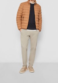 Giacca puffer color sabbia con design a righe orizzontali, indossata sopra un maglione scuro, abbinata a pantaloni beige chiaro e scarpe color sabbia.