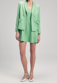 Mátově zelený blazer s dvojitými knoflíky, zkombinovaný s odpovídajícími šaty. Model má na sobě bílé sandály na podpatku s dekorativními prvky.