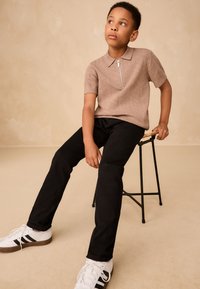 Polo bej cu mâneci scurte și model geometric texturat, cu guler cu fermoar, asortat cu pantaloni negri și adidași albi cu dungi negre.