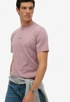 Mężczyzna w mauve krótkim rękawie t-shirtu, niebieskich dżinsach, szarej bluzie zawiązanej w pasie i beżowym zegarku na nadgarstku, spoglądający przez lewe ramię.