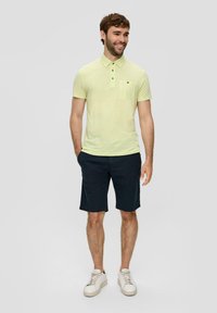 Svijetložuta polo majica s ovratnikom i kratkim rukavima, u kombinaciji s tamnoplavim šortsevima. Model nosi bijele tenisice, stoji opušteno.