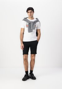 Camiseta gráfica blanca con un estampado colorido, pantalones cortos negros enrollados, suéter gris colgado sobre los hombros, zapatos negros con suelas gruesas.