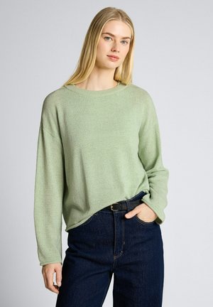 Frau mit langen blonden Haaren, die einen hellgrünen Pullover und dunkelblaue Jeans trägt, posiert mit einer Hand in der Tasche vor einem schlichten Hintergrund.