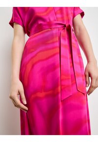 Długa różowa sukienka z wiązaniem w talii, o płynnej konstrukcji z pomarańczowymi i ciemnoróżowymi falistymi wzorami, wykonana z gładkiego, błyszczącego materiału.