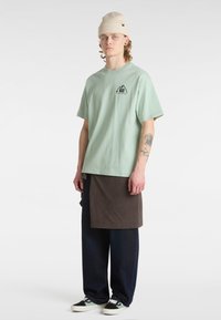 Camiseta de algodón verde menta con un logo de montaña, combinada con una falda marrón, acompañada de pantalones de denim oscuro y zapatillas negras con acentos verdes.