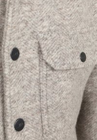 Lichtbeige wollen jas met een herringbone textuur, zwarte knopen en een klepzak aan de linkerkant.