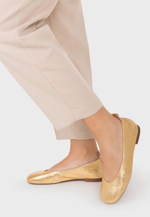 Pies con bailarinas doradas brillantes combinadas con pantalones cortos beige sobre un fondo claro.