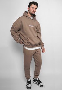 Brauner oversized Hoodie mit weißem Logo, kombiniert mit passenden Jogginghosen. Das Material scheint weich zu sein; beide Teile haben eine lockere Passform. Schwarz-weiße Sneaker.