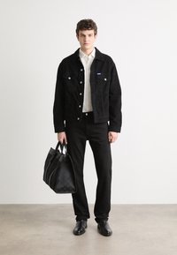 Veste en velours côtelé noir avec des boutons blancs, associée à une chemise blanche et un pantalon noir. Le modèle tient un sac fourre-tout noir texturé. Chaussures noires.
