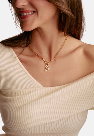 Goldene Halskette mit verlängerten Gliedern, verziert mit einem Herz- und Schlüsselanhänger sowie einem weißen Perlenakzent. Getragen über einem cremefarbenen, gerippten Oberteil.