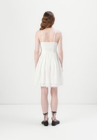 Weißes Spitzenkleid mit eng anliegendem Oberteil, weit ausgestelltem Rock und verstellbaren Trägern. Kombiniert mit schwarzen Schnürschuhen. Sichtbare strukturierte Stoffdetails.