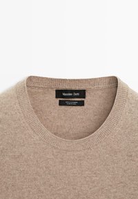 Beige kashmirtröja med rund halsringning, ribbade muddar och fåll. Inuti etiketten finns varumärke och materialinformation.