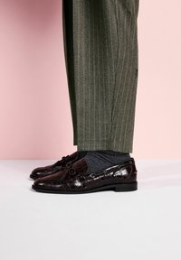 Mocasini maro din piele cu aspect crocodil, cu accente de șireturi, asortați cu pantaloni gri cu model și șosete închise la culoare, pe un fundal roz.