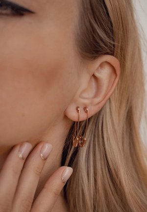 Rosenvergoldete Ohrhänger mit verlängerten Haken und ovalen Enden, die an dem Ohr eines Modells hängen. Das Modell hat helles, strukturiertes Haar und manikürte Nägel.