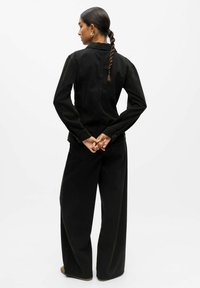 Giacca di jeans nera con silhouette aderente, chiusure con bottoni e maniche lunghe. Indossata con pantaloni neri a gamba larga, vista da dietro.